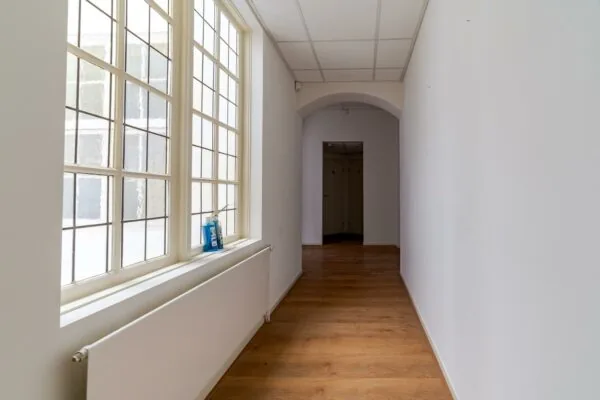 Lichte gang met houten vloer, grote ramen aan de linkerkant en een nis aan het einde in een gebouw aan het Lange Voorhout.